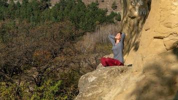 mujer con los ojos cerrados y los brazos detrás de la cabeza sentada en una montaña en posición de loto en meditación en un día de otoño foto