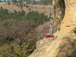 mujer con los ojos cerrados y los brazos levantados sentada en una montaña en posición de loto en meditación en un día de otoño foto