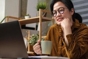 mujer asiática bebe una taza de café y ve la transmisión en vivo en una computadora portátil cuando trabaja en casa foto