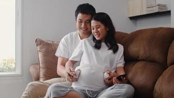 Young Asian Pregnant couple using phone and headphone play music for baby in belly. Mom and Dad feeling happy smiling peaceful while take care child lying on sofa in living room at home concept. photo