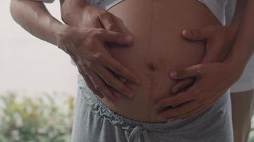 Young Asian Pregnant couple hug and holding belly talking with their child. Mom and Dad feeling happy smiling peaceful while take care baby, pregnancy near window in living room at home concept. photo