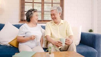 Asian elderly couple drinking warm coffee and talking together in living room at home, couple enjoy love moment while lying on sofa when relaxed at home. Lifestyle senior family at home concept. photo