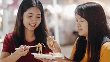 Asian young women friends travel in Bangkok, Thailand, beautiful female feeling happy walking and eating Pad Thai at The Khao San Road. Women travel eat street food in Thailand concept.