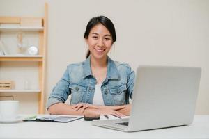 hermosa joven sonriente mujer asiática trabajando con ordenador portátil en el escritorio en la sala de estar en casa. Mujer de negocios de Asia escribiendo finanzas de documentos de cuaderno y calculadora en la oficina en casa. disfrutando del tiempo en el concepto de casa. foto