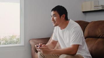 Young Asian man using joystick playing video games in television in living room, male feeling happy using relax time lying on sofa at home. Men play games relax at home concept. photo