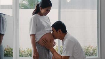 joven pareja asiática embarazada besando a su esposa en el vientre hablando con su hijo. mamá y papá se sienten felices sonriendo en paz mientras cuidan al bebé, el embarazo cerca de la ventana en la sala de estar en el concepto de hogar. foto
