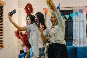 Group of Asian women party at home, female using mobile phone making vlog and selfie to social media while funny moment dancing together in living room in night. Young friend celebrate holiday concept photo