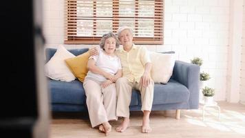 Asian elderly couple watching television in living room at home, sweet couple enjoy love moment while lying on the sofa when relaxed at home. Enjoying time lifestyle senior family at home concept. photo