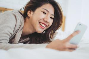 Happy Asian women are using smart phone on the bed in morning. Asian woman in bed checking social apps with smartphone. Smiling woman surfing net with cellphone at home. Mobile addict concept. photo