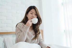 hermosa mujer asiática disfrutando de un café caliente en la cama en su dormitorio. relajación en la cama. hermosa mujer asiática con suéter cómodo sosteniendo una taza de café. estilo de vida mujer asiática en el concepto de hogar. foto