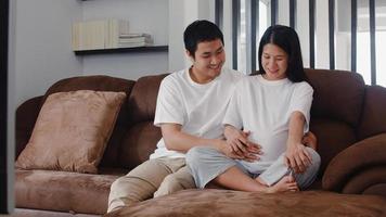 Young Asian Pregnant couple man touch his wife belly talking with his child. Mom and Dad feeling happy smiling peaceful while take care baby, pregnancy lying on sofa in living room at home concept. photo