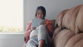 Young Asian Pregnant woman using tablet search pregnancy information. Mom feeling happy smiling positive and peaceful while take care her child lying on sofa in living room at home concept. photo