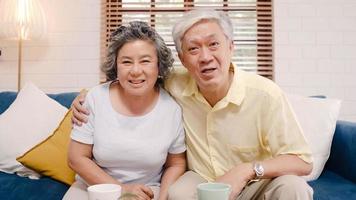 Asian elderly couple using smartphone video conference with grandchild while lying on sofa in living room at home. Enjoying time lifestyle senior family at home concept. Portrait looking at camera. photo