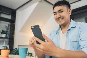 Young Asian man using smartphone while lying on the desk in her living room. Happy male use phone for texting, reading, messaging and buying online at home. Lifestyle man at home concept. photo