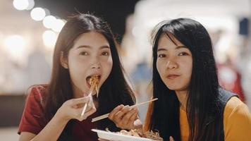 Asian young women friends travel in Bangkok, Thailand, beautiful female feeling happy walking and eating Pad Thai at The Khao San Road. Women travel eat street food in Thailand concept. photo