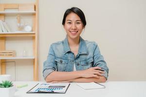 mujer de negocios de asia que se siente feliz sonriendo y mirando a la cámara mientras se relaja en la oficina en casa. joven asiática trabajando escribiendo finanzas de documentos de cuaderno en el escritorio en la sala de estar en casa. foto