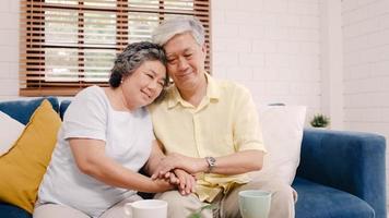 Asian elderly couple holding their hands while taking together in living room, couple feeling happy share and support each other lying on sofa at home. Lifestyle Senior family at home concept. photo