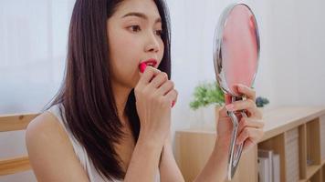 Young Asian woman using lipstick make up in front mirror, Happy female using beauty cosmetics to improve herself ready to working in bedroom at home. Lifestyle women at home concept.