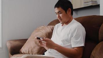 Young Asian man using mobile phone playing video games in television in living room, male feeling happy using relax time lying on sofa at home. Men play games relax at home concept. photo