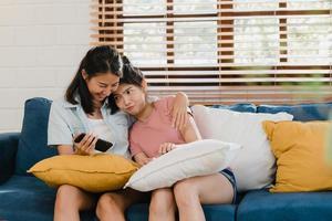 Young Lesbian lgbtq women couple using mobile phone at home. Asian lover female happy relax laugh and fun using technology play games in internet together while lying sofa in living room concept. photo