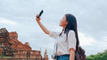 Young Asian backpacker blogger woman casual take selfie video call on mobile phone sharing live online for audience travel together at pagoda old city, Lifestyle tourist travel holiday concept. photo