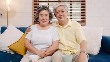 Asian elderly couple feeling happy smiling and looking to camera while relax on the sofa in living room at home. Enjoying time lifestyle senior family at home concept. Portrait looking at camera. photo