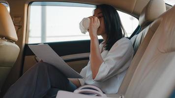 Successful young Asia businesswoman in fashion office clothes working and drinking disposable paper cup of hot drink in back seat of car in urban modern city in morning. Business on the go concept. photo