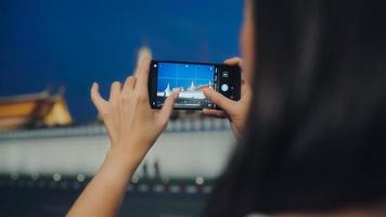 feliz joven turista de asia con estilo casual mantenga el teléfono móvil relájese alegre tome una foto pacífica del templo en la ciudad de bangkok por la noche. concepto de vacaciones de viajes turísticos de estilo de vida.