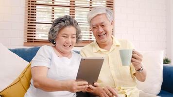 Asian elderly couple using tablet and drinking coffee in living room at home, couple enjoy love moment while lying on sofa when relaxed at home. Enjoying time lifestyle senior family at home concept. photo