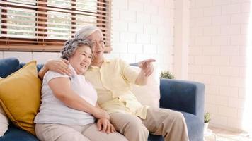 Asian elderly couple watching television in living room at home, sweet couple enjoy love moment while lying on the sofa when relaxed at home. Enjoying time lifestyle senior family at home concept. photo