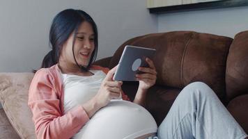 Young Asian Pregnant woman using tablet search pregnancy information. Mom feeling happy smiling positive and peaceful while take care her child lying on sofa in living room at home concept. photo