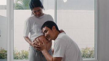 joven pareja asiática embarazada besando a su esposa en el vientre hablando con su hijo. mamá y papá se sienten felices sonriendo en paz mientras cuidan al bebé, el embarazo cerca de la ventana en la sala de estar en el concepto de hogar. foto