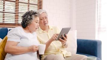 Asian elderly couple using tablet and drinking coffee in living room at home, couple enjoy love moment while lying on sofa when relaxed at home. Enjoying time lifestyle senior family at home concept. photo