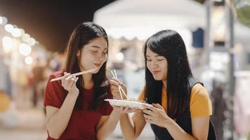 Asian young women friends travel in Bangkok, Thailand, beautiful female feeling happy walking and eating Pad Thai at The Khao San Road. Women travel eat street food in Thailand concept. photo