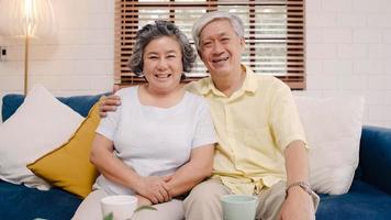 Asian elderly couple feeling happy smiling and looking to camera while relax on the sofa in living room at home. Enjoying time lifestyle senior family at home concept. Portrait looking at camera. photo