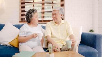 Asian elderly couple drinking warm coffee and talking together in living room at home, couple enjoy love moment while lying on sofa when relaxed at home. Lifestyle senior family at home concept. photo