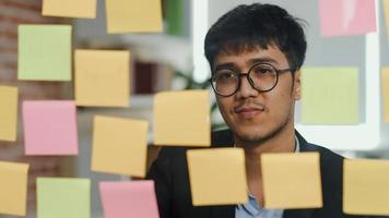 un hombre creativo asiático pega una nota adhesiva en una placa de vidrio. joven hombre de negocios profesional escribe información, estrategia, recordatorio en papel, situación comercial, puesta en marcha en el concepto de oficina de loft. foto