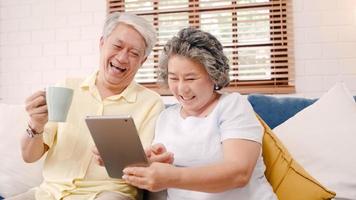 Asian elderly couple using tablet and drinking coffee in living room at home, couple enjoy love moment while lying on sofa when relaxed at home. Enjoying time lifestyle senior family at home concept. photo
