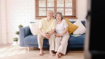 Asian elderly couple watching television in living room at home, sweet couple enjoy love moment while lying on the sofa when relaxed at home. Enjoying time lifestyle senior family at home concept. photo