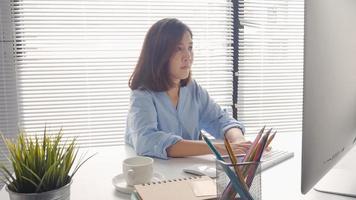 Beautiful young smiling Asian woman working on laptop while at home in office work space. Businesswoman working from home via portable computer writing on keyboard. Enjoying time at home. photo