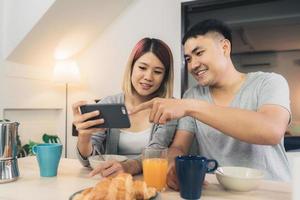 Atractiva pareja asiática joven distraída en la mesa con el periódico y el teléfono celular mientras desayuna. Emocionada joven pareja asiática sorprendida por una increíble buena noticia, familia feliz asombrada por Internet. foto