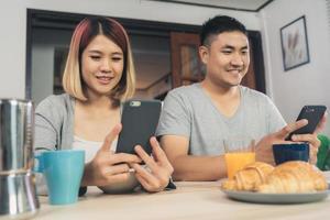 Atractiva pareja asiática joven distraída en la mesa con el periódico y el teléfono celular mientras desayuna. Emocionada joven pareja asiática sorprendida por una increíble buena noticia, familia feliz asombrada por Internet. foto