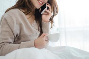 mujer asiática usando el teléfono inteligente en su cama mientras sostenía una taza de café por la mañana. hermosa mujer asiática disfrutando de un café caliente y hablando por teléfono en su dormitorio. concepto de mujer asiática de estilo de vida foto