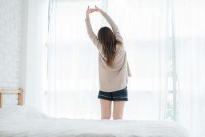 Back view of happy beautiful young Asian woman waking up in morning, sitting on bed, stretching in cozy bedroom, looking through window. Funny woman after wake up. She is stretching and smiling. photo