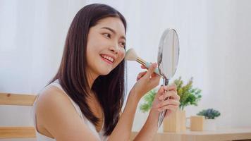 Asian woman using powder by brush make up in front mirror, Happy female using beauty cosmetics to improve herself ready to working in bedroom at home. Lifestyle women at home concept.