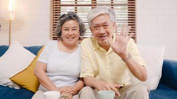 Asian elderly couple using smartphone video conference with grandchild while lying on sofa in living room at home. Enjoying time lifestyle senior family at home concept. Portrait looking at camera. photo