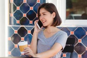 sonrisa feliz mujeres de negocios asiáticas usando un teléfono celular parlante sentado en un café y sosteniendo una taza de café. hermosa mujer asiática alegre en el café hablando por teléfono y sonriendo mientras disfruta del café. foto