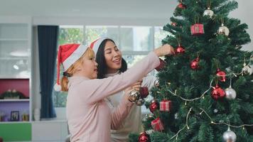 amigas asiáticas decoran el árbol de navidad en el festival de navidad. mujer adolescente feliz sonriendo celebrar las vacaciones de invierno de Navidad juntos en la sala de estar en casa.