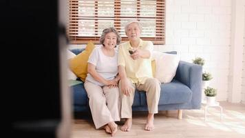 Asian elderly couple watching television in living room at home, sweet couple enjoy love moment while lying on the sofa when relaxed at home. Enjoying time lifestyle senior family at home concept. photo
