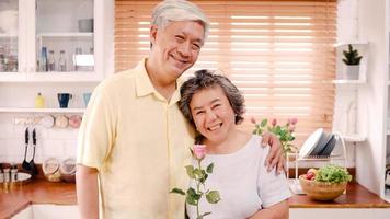 Asian elderly couple feeling happy smiling and holding flower and looking to camera while relax in kitchen at home. Lifestyle Senior family enjoy time at home concept. Portrait looking at camera. photo